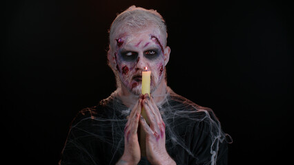 Frightening man with Halloween zombie bloody wounded makeup spells conjures over a candle. Horror theme. Sinister undead guy isolated on studio black background. Voodoo rituals. Fashion body art