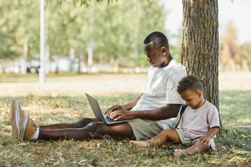 Side view portrait of young African-American man using laptop outdoors in park with cute baby son, copy space