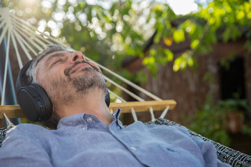 Homme d'âge mur 50 ans se repose dans son hamac en écoutant de la musique avec un casque à l'ombre d'un arbre. Il profite de la vie - obrazy, fototapety, plakaty