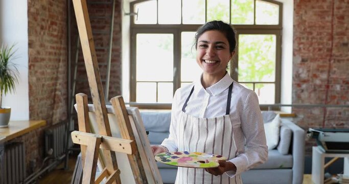 Young 25s attractive Indian woman wear apron holding palette and paintbrush, standing near easel posing alone in art school space smiling looking at camera. Artistic studio, creativity, hobby concept