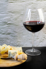 wooden tray with grapes and various varieties of soft and hard cheese and a glass of red wine against the background of a white brick wall