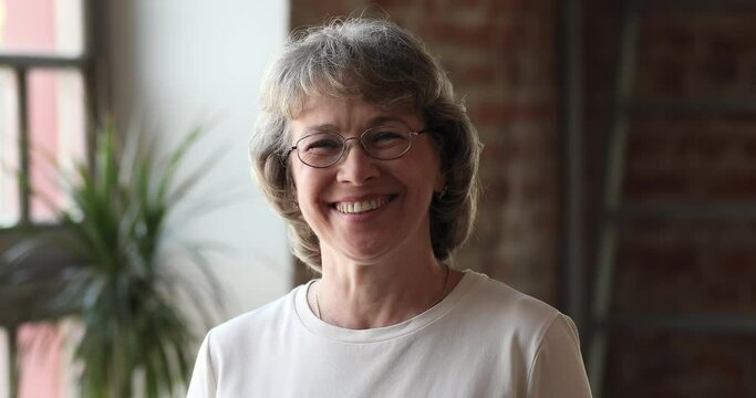 Head shot portrait older grey haired 60s woman in glasses standing indoor smile look at camera. Dental and vision eyesight check up services for older citizen, professional occupation person concept