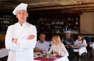 Portrait of confident professional pizzeria chef in restaurant hall on background with guests - obrazy, fototapety, plakaty