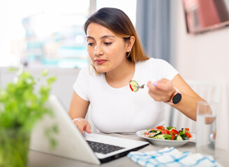 Positive young latino woman eating salad and chatting online using laptop