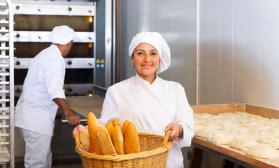 Portrait of successful hispanic female baker holding wicker basket with fresh baked baguettes in bakery