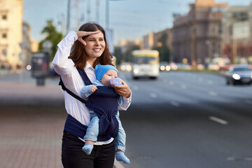 Mother with little boy in sling stands near traffic on street.