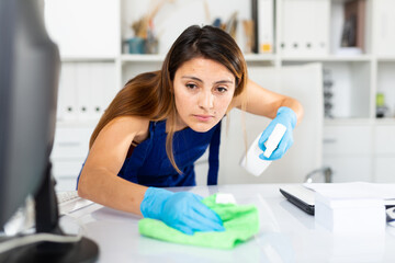 Hispanic woman in uniform and gloves cleaning desk in modern business office