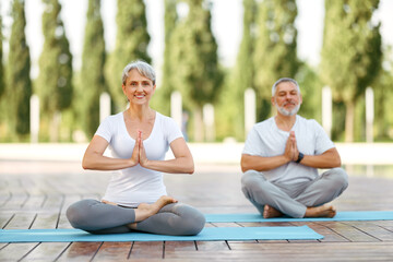 Fototapeta na wymiar Happy senior husband and wife meditating in lotus position during yoga outdoors
