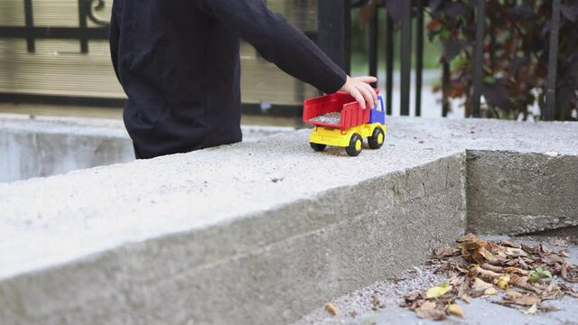 caucasian baby kid boy in black long sleeve shirt plays with colorful plastic red blue yellow toy car truck outside in warm day autumn weather. child drives vehicle on grey cement road with one hand