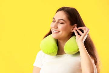 Beautiful young woman with travel pillow putting ear plug on yellow background