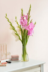 Vase with beautiful gladiolus flowers, female accessories and cosmetics on table against color wall