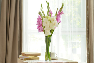 Vase with beautiful gladiolus flowers, books and cup of coffee on table near window