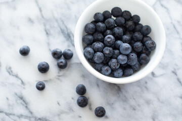 An overhead view of a white bowl of blueberries next to loose blueberries on a grey marble background.