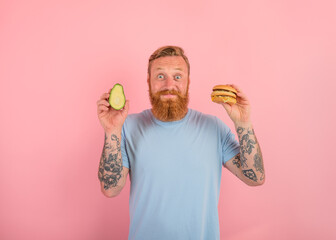Happy man with beard and tattoos is undecided if to eat an avocado or an hamburger