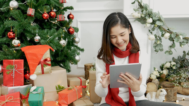 Asian woman using smart tablet sit at christmas tree xmas present gift box in cozy living room. Woman hand touch screen smile shopping online, happiness time christmas holiday winter season festive