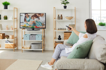 Woman watching television sitting on couch at home. Happy relaxed lady resting on sofa in living room and switching on TV set with big black empty blank screen to watch show on her favorite channel