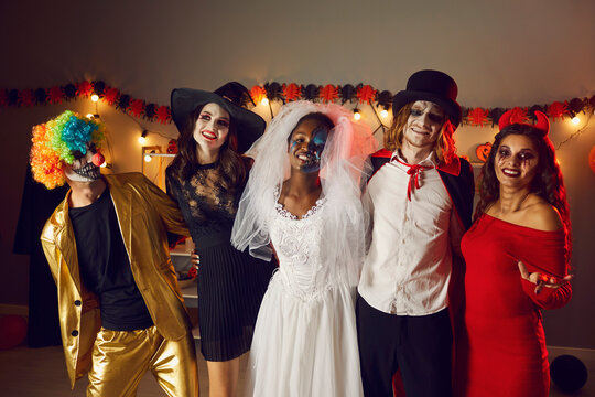 Portrait of multiracial adult friends in spooky costumes for Halloween at home party. People in images of clown, vampire, witch, dead bride and devil woman hug and smile looking at camera in dark room
