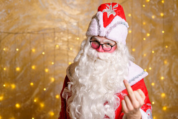 Portrait of an angry santa claus wearing glasses with white long beard showing middle finger marks against a wall background with garland. A man in costume of a fairytale hero shows a finger gesture