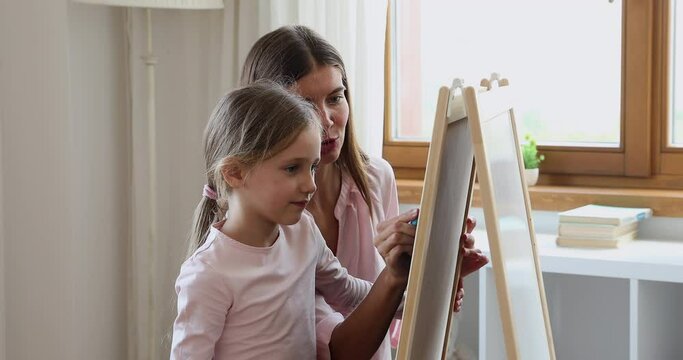 Smiling young mother give art lesson to little girl stand by drawing board hold colored chalk show teach sketching basics. Small daughter imitate mom create paint picture on whiteboard using crayons