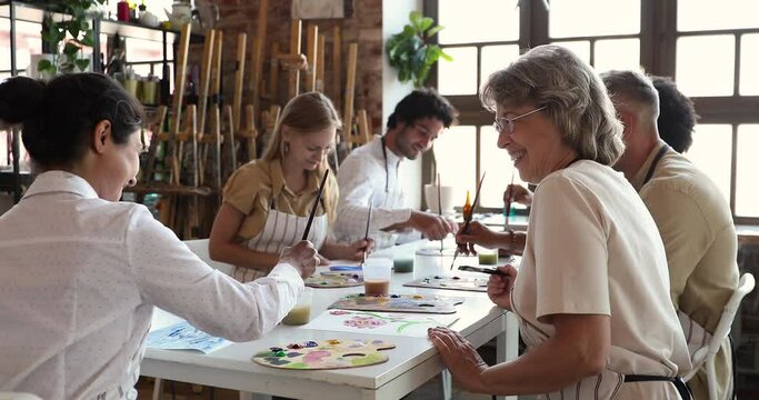 Art therapy, creative group class in artistic studio concept. In modern workshop elderly 60s young 30s women participate painting lesson sit at table talking laughing enjoy process and communication