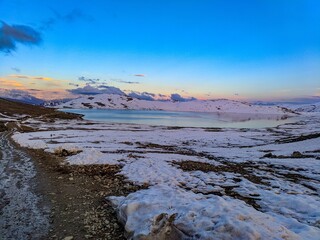 Ice lake in snow covered ground with small stones placed on the ground with orange clouds in blue sky and hills covered with snow