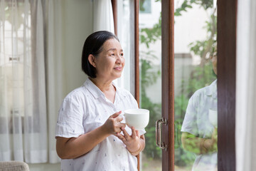 Senior elderly Asian woman drinking tea or coffee near the windows at home. Retirement, health care and ageing concept