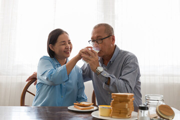 Asian elderly couple having bread breakfast and drinking milk with carefully in the kitchen room at home