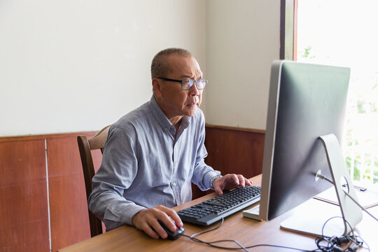 Asian senior businessman wearing glasses and sitting working with desktop computer at home. Asian elderly man using desktop computer in the workplace
