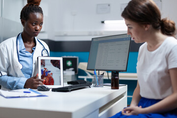 African american cardiologist doctor holding tablet explaining heart xray discussing medical expertise with patient woman during clinical appointment. Radiologist working in hospital office.
