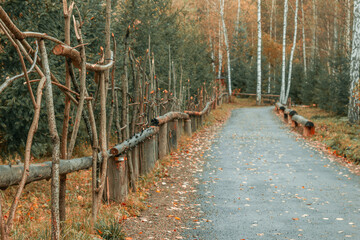 Fototapeta premium A view of the road along a beautiful fence made of branches with fallen leaves against the backdrop of an autumn forest. The road in the park in autumn