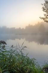 Sunrise over the foggy lake with the reflection of sun. Mist on the water, forest silhouettes and the rays of the rising sun. Beautiful morning landscape with sunrise over river.