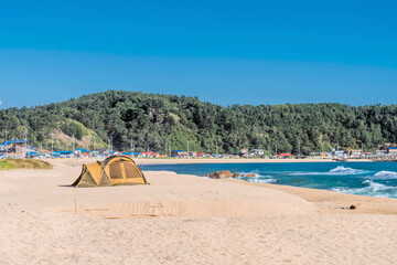 Brown tent pitched on sandy ocean beach with village buildings in background.