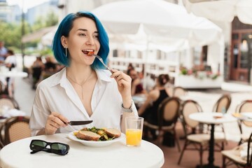Stylish young woman eating healthy salad on a restaurant terrace, feeling happy on a summer day