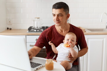 Positive optimistic handsome freelancer male wearing burgundy t shirt, posing in white kitchen, sitting in front of laptop with baby in hands and working online.