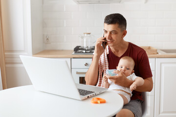 Indoor shot of man wearing burgundy casual t shirt with towel on his shoulder, looking after baby...