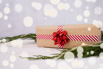 Christmas gift made of kraft paper with a red bow on a background of coniferous branches of blurry lights.