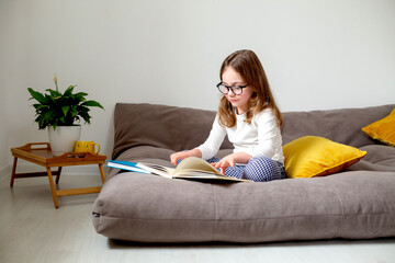 Obraz premium a cute little girl in glasses, jeans and a white turtleneck is reading a book, doing homework, preparing for exams sitting on the bed. home education. space for text. High quality photo