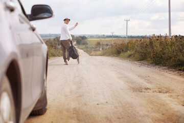 Obraz premium A man walks along a country road. Hitchhiker around the country. A man stops a passing car on the road.