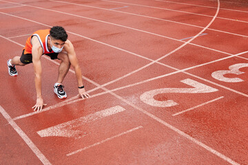 Young latin guy with protective mask, in position to run at full speed on a track, ready, at the start line, copy space