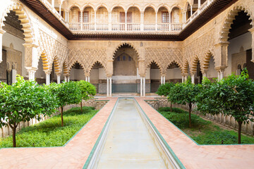 Obraz premium Water basin in a beautiful patio with green plants and arab style decorated arches in the famous Alcazar (meaning: fortress), Seville, Andalusia, Spain.