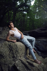 Naklejka na ściany i meble sexy trendy teen guy in a white T-shirt and blue jeans is sitting on a rock by a waterfall
