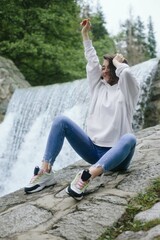 Woman traveler sitting on a rock in front of a waterfall high in the mountains, space for text, atmospheric epic moment