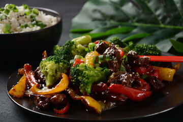 Asian teriyaki beef with red and yellow bell peppers, broccoli and sesame seeds on a plate on the table. Spicy teriyaki beef stir fry with vegetables and rice  on a dark background