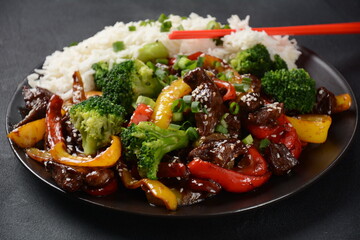 Asian teriyaki beef with red and yellow bell peppers, broccoli and sesame seeds on a plate on the table. Spicy teriyaki beef stir fry with vegetables and rice  on a dark background