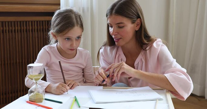 Drawing game. Young adult woman mum babysitter sit at table with little preteen girl help kid in creating picture using colored pencils. Millennial mom assist child schoolgirl in making sketch on time
