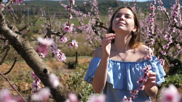 Positive european girl in blue dress standing under blossom peach tree at sunny spring day