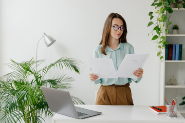 Young successful employee business woman in blue shirt hold paper account documents work stand at workplace white desk with laptop pc computer at light modern office indoor Achievement career concept