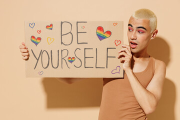 Young amazed fun friendly blond latin gay man with make up in beige tank shirt hold card sign with...