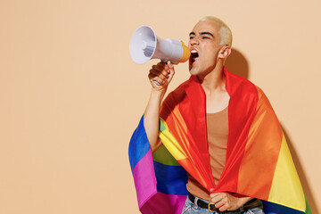 Young blond latin gay man with make up in beige tank shirt wrapped in rainbow flag hold scream...