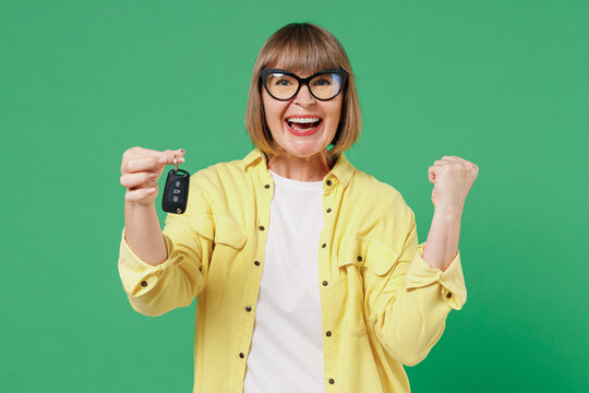 Elderly Smiling Happy Jubilant Cool Woman 50s In Glasses Yellow Shirt Hold Car Keys Fob Keyless System Do Winner Gesture Isolated On Plain Green Background Studio Portrait People Lifestyle Concept